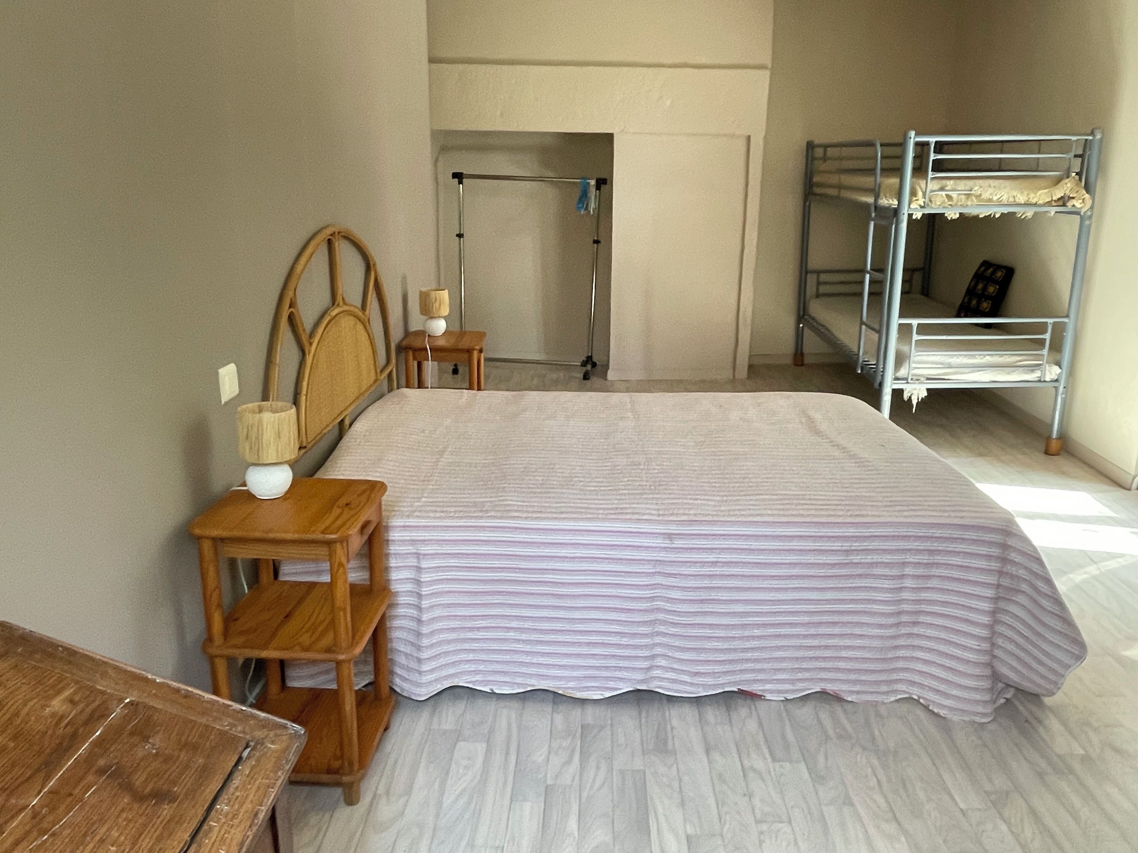 Un dormitorio con cama, mesitas de noche de madera, cabecero de madera, literas metálicas y una puerta que da a otra habitación.