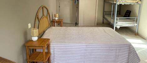 Un dormitorio con cama, mesitas de noche de madera, cabecero de madera, literas metálicas y una puerta que da a otra habitación.