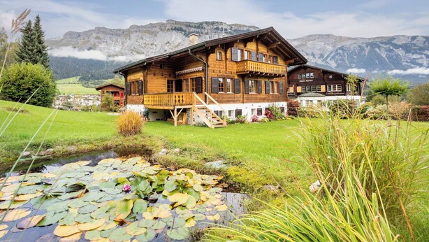 A wooden chalet with a balcony, a pond with lily pads, and a mountainous backdrop.
