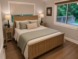 A bedroom with a wooden bed, two bedside tables with lamps, a window with a view of greenery, and a framed picture on the wall.