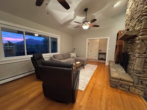 A living room with a stone fireplace, a ceiling fan, and a large window overlooking a scenic view.