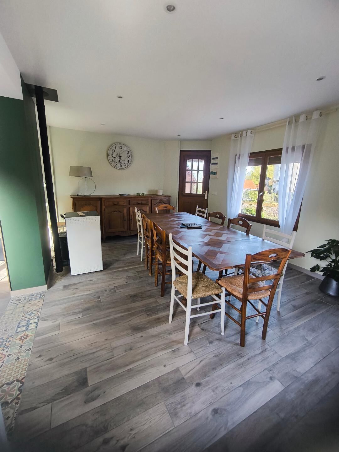 Een eetgedeelte met een houten tafel en stoelen, een klok aan de muur en een raam met gordijnen.