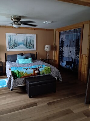 A bedroom with a bed, a ceiling fan, a painting, a lamp, and a deer-themed bedspread.