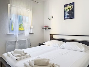 Ein Hotelzimmer mit einem Bett, weißer Bettwäsche, einem Fenster mit durchscheinenden Vorhängen, einem Wandbild und einem Nachttisch mit einer Vase.