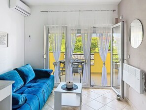 Ein Wohnzimmer mit einem blauen Sofa, einem kleinen Tisch, einem Heizkörper und einer Schiebetür zu einem Außenbereich mit Tisch und Stühlen.