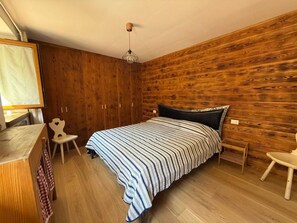 Ein Schlafzimmer mit Holzwand, einem Bett mit gestreifter Bettdecke, einem Sessel, einem Schreibtisch und einer Hängelampe.