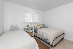 Quarto com duas camas, uma mesa de cabeceira branca, janela com persianas e uma prateleira pequena com objetos decorativos.