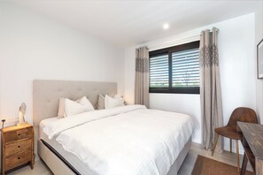 Ein Schlafzimmer mit einem großen Bett, einem Nachttisch mit Lampe, einem Sessel und einem Fenster mit Vorhängen.