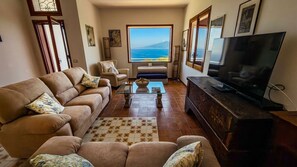 Ein Wohnzimmer mit Sofa, Sessel, Couchtisch und Fernseher, mit Blick auf das Meer durch große Fenster.