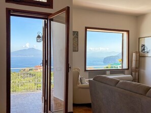 Ein Zimmer mit großem Fenster, das Blick auf Berg und Meer gewährt, einer Couch und einer Tür zu einem Balkon.