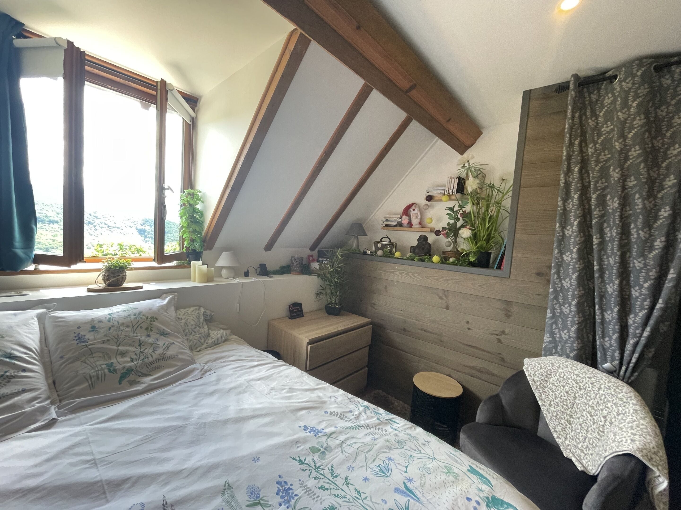 Un dormitorio con techo inclinado, una cama con ropa de cama floral, una mesita de noche, una lámpara, un estante pequeño con libros y objetos decorativos, y una ventana con cortinas.
