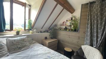 Un dormitorio con techo inclinado, una cama con ropa de cama floral, una mesita de noche, una lámpara, un estante pequeño con libros y objetos decorativos, y una ventana con cortinas.