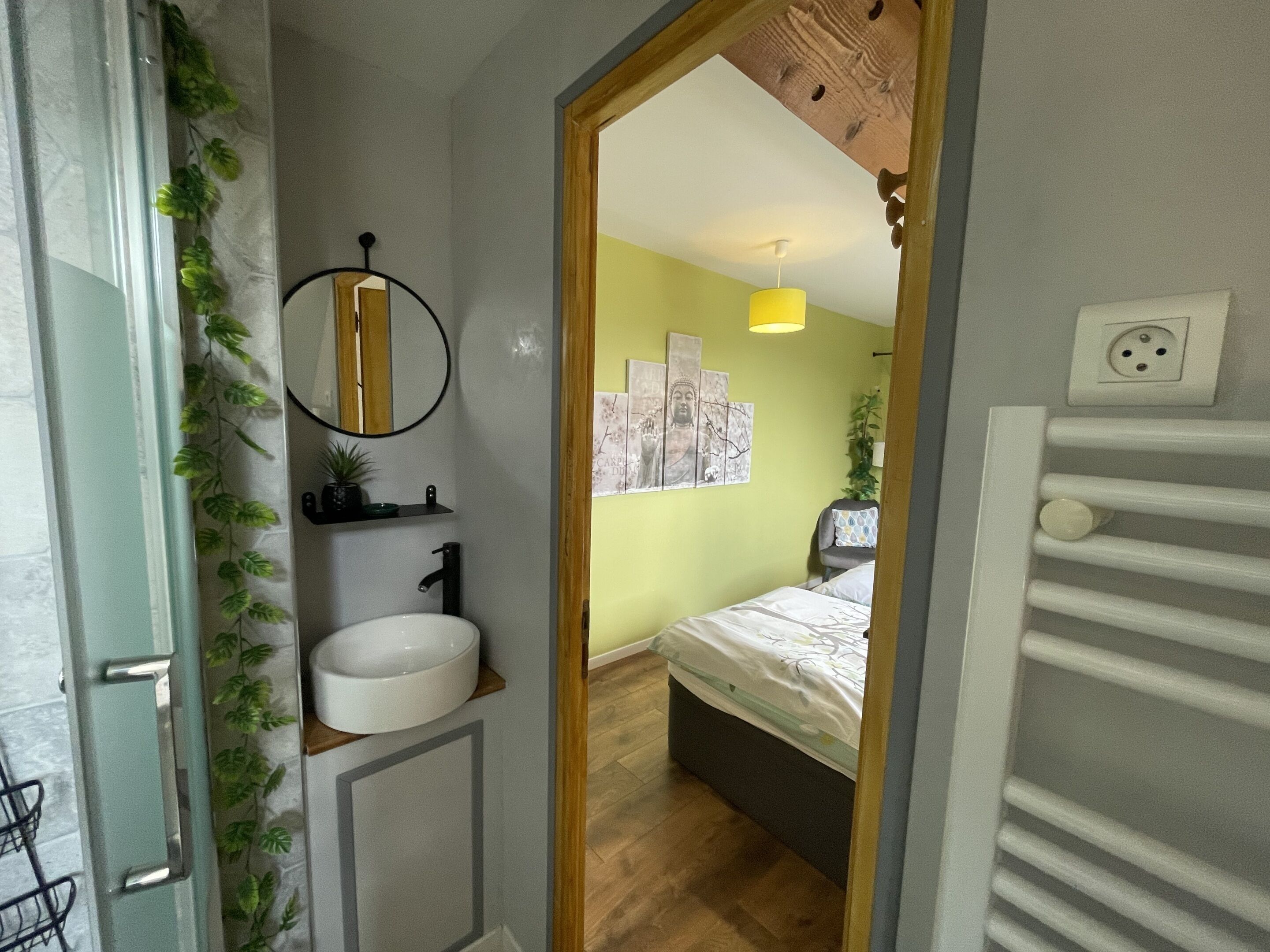 A narrow hallway leading to a bedroom with a bed, a bedside table, a mirror, a wall-mounted shelf with a plant, and a light fixture.