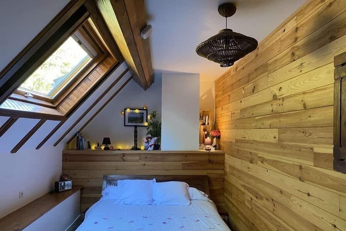 Un dormitorio con techo inclinado, paredes de madera, una cama con ropa de cama blanca, una mesita de noche pequeña, una lámpara y un marco de fotos en la pared.