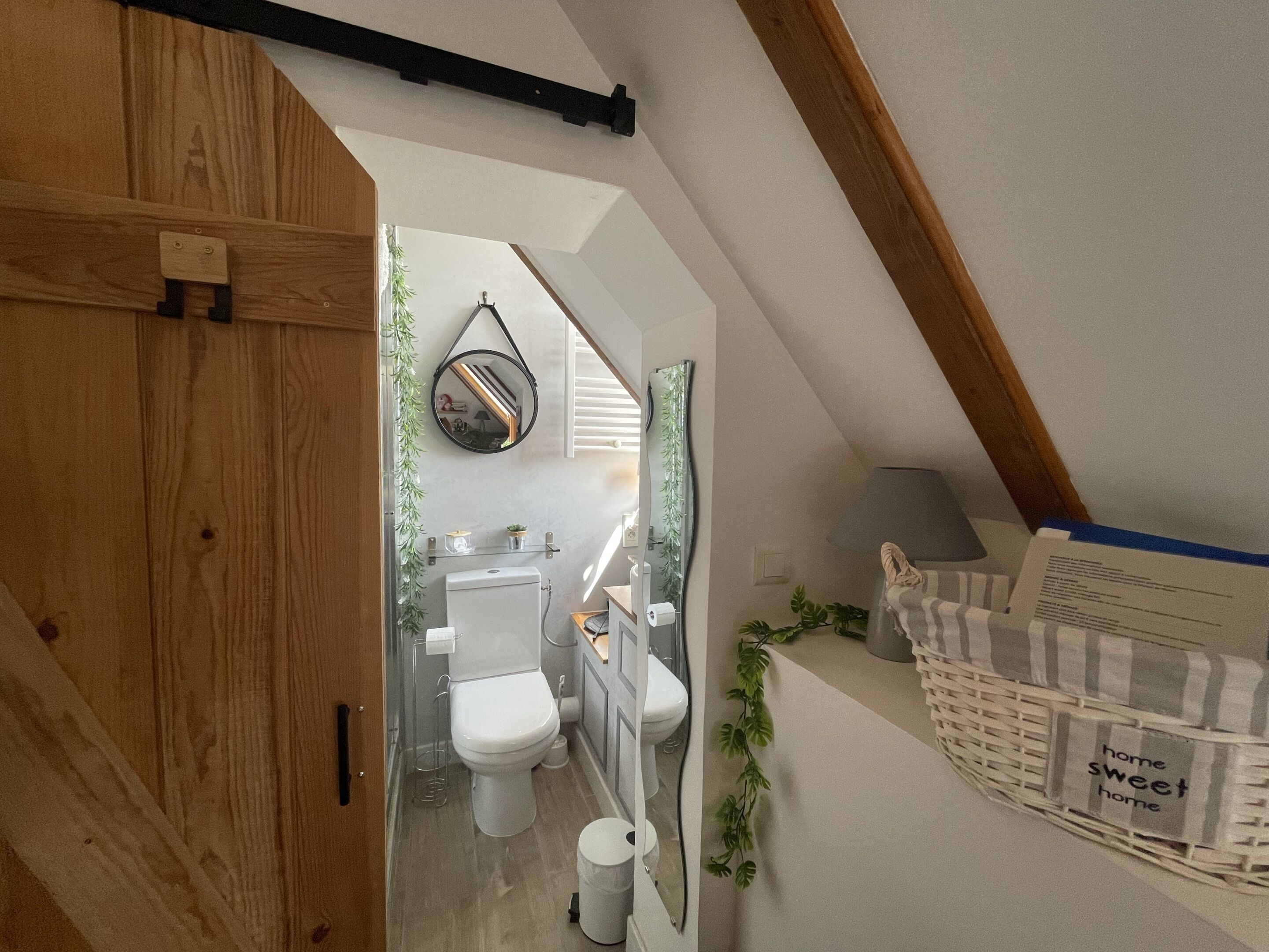 Un dormitorio abuhardinado con estructura de madera, techo inclinado, una ventana pequeña y una cesta de mimbre blanca con una etiqueta.