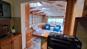 A kitchen with wooden cabinets, a sink, and a stove. There is a dining area with a table and chairs, and a view of the outside. - Anca on C With Seaview (Klein Brak River)