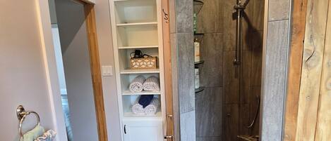 Un pasillo estrecho con suelo de madera, un armario blanco con estantes y un área de ducha con suelo de azulejos.