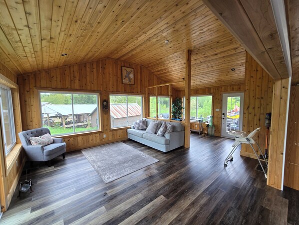 A wooden cabin interior with a sofa, armchair, and large windows offering views of the outdoors.