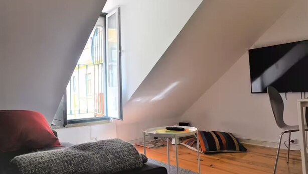 Une pièce avec un plafond en pente, un téléviseur fixé au mur, une petite table avec un coussin et une fenêtre avec des stores.