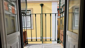 Une vue à travers une porte vitrée, avec un portail en métal, qui donne sur un mur jaune et une fenêtre avec des volets.