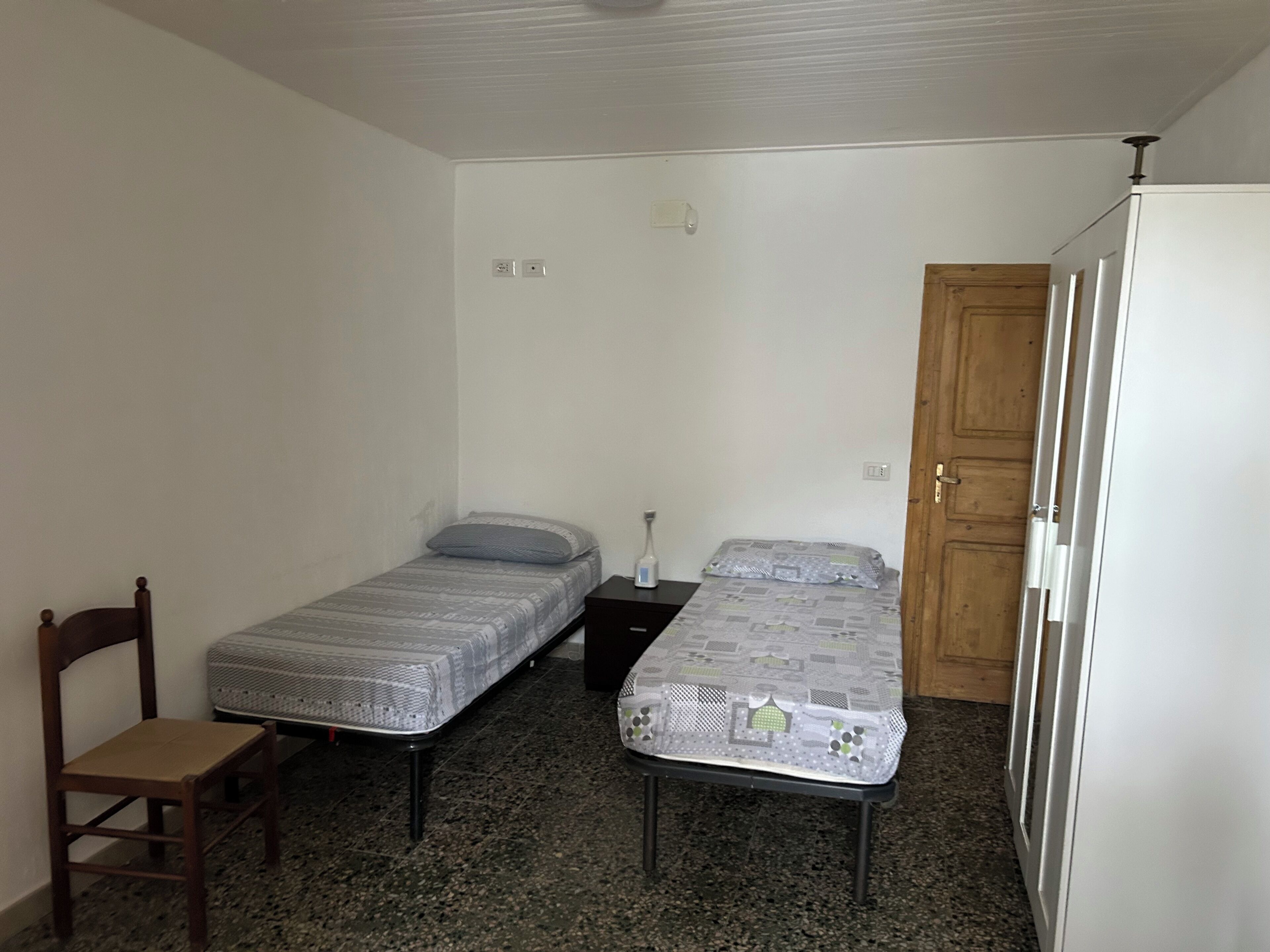 A small room with two single beds, a wooden chair, a white refrigerator, and a wooden door.