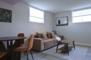 A living room with a sofa, a coffee table, a dining table with chairs, and two windows.