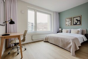 Ein modernes Schlafzimmer mit großem Fenster, einem Schreibtisch mit Stuhl, einem Bett mit weißer Bettdecke und zwei abstrakten Wandbildern.