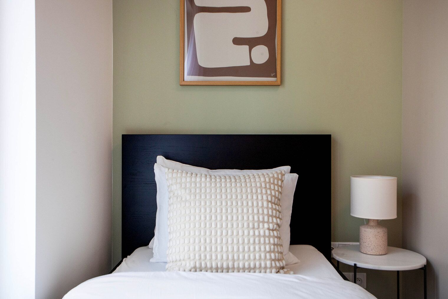 Cama bem arrumada com travesseiro branco e cabeceira escura. Uma pequena mesa com abajur fica ao lado da cama.