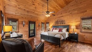 A wooden cabin bedroom with a bed, two bedside tables, a chair, a ceiling fan, and a window. - Grounding Lodge | Sauna + Cold Plunge + Hot Tub (Blue Ridge)