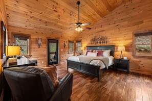 Ein Schlafzimmer einer Holzhütte mit einem Bett, zwei Nachttischen, einem Sessel, einem Deckenventilator und einem Fenster.