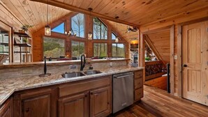 A wooden cabin interior with a kitchen featuring granite countertops, a dishwasher, and a large window offering a view of trees. - Grounding Lodge | Sauna + Cold Plunge + Hot Tub (Blue Ridge)