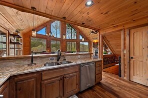 Ein Holzblockhaus-Interieur mit einer Küche, die sich durch Granit Arbeitsplatten, einen Geschirrspüler und ein großes Fenster mit Blick auf Bäume auszeichnet.