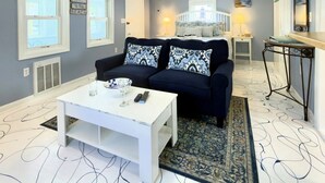 A room with a dark blue sofa, a white coffee table, a patterned rug, a bed with white bedding, and a small table with a lamp.