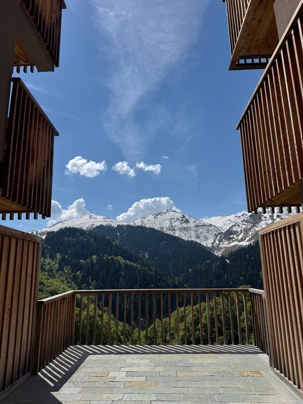 Ein Balkon mit Steinboden, Holzgeländern und Blick auf schneebedeckte Berge und bewaldete Hügel.