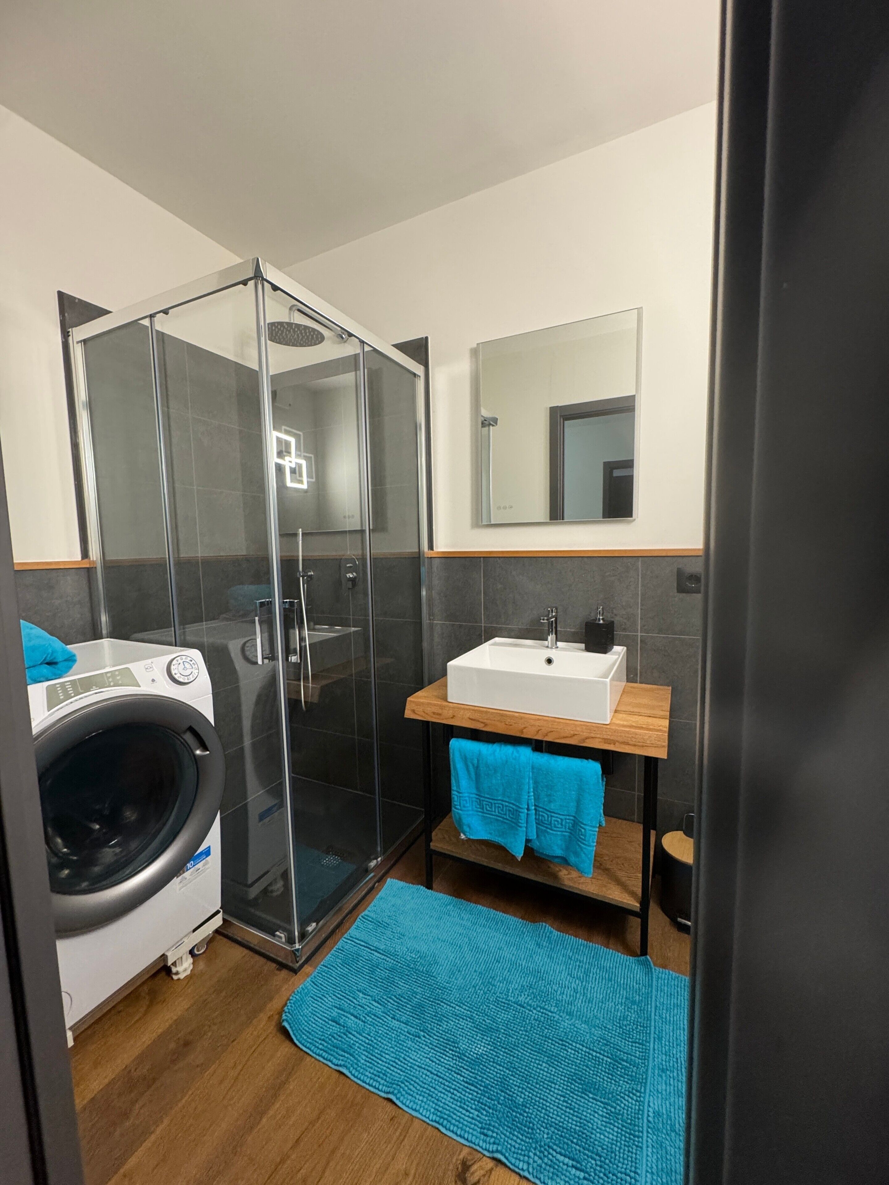 Ein modernes Badezimmer mit Duschkabine aus Glas, einer Waschmaschine, einem Waschbecken mit Spiegel und einem blauen Handtuch auf einem Holzregal.