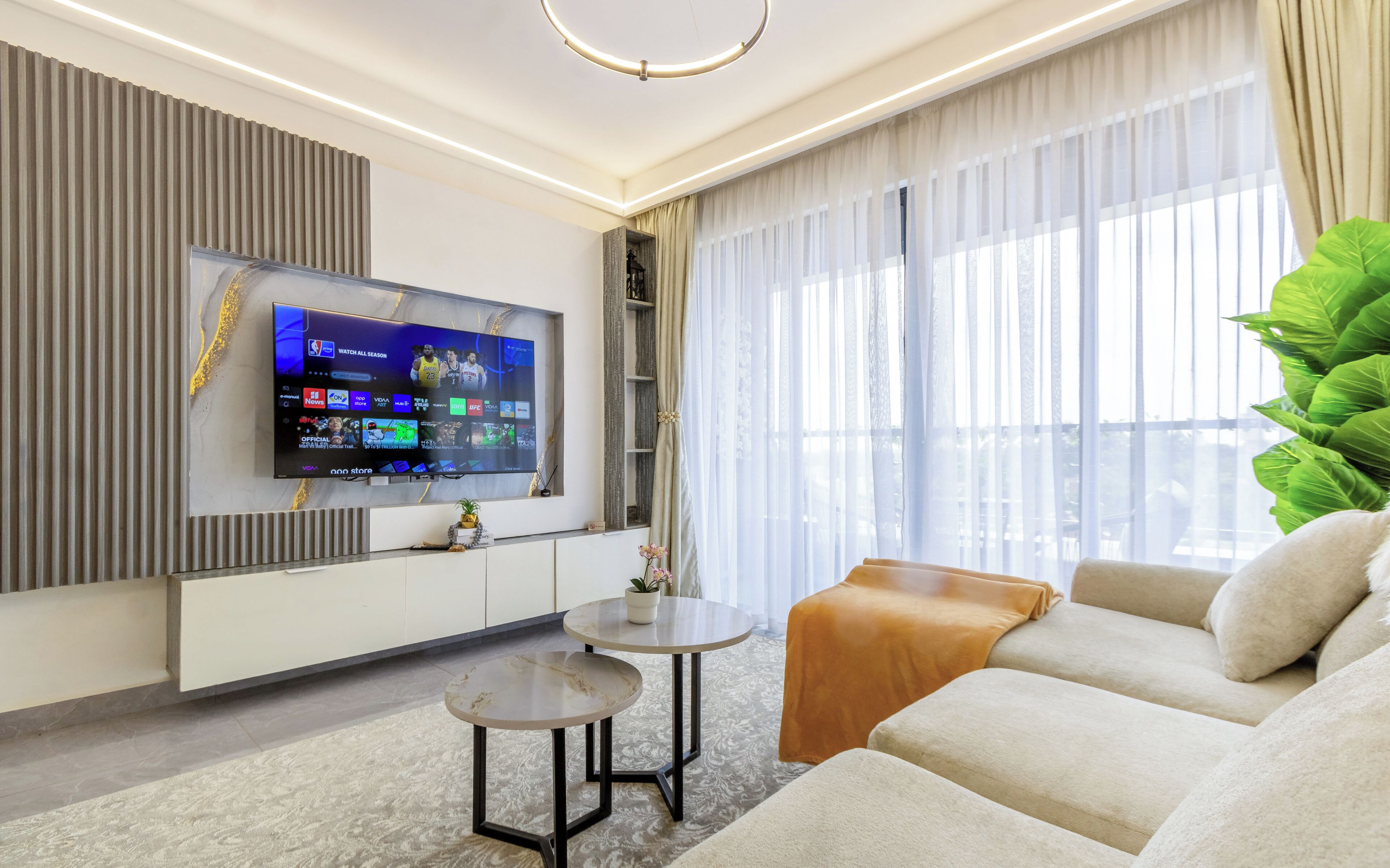 Un soggiorno moderno con una TV a schermo piatto, un divano, un tavolino da caffè e un'ampia finestra con tende leggere.