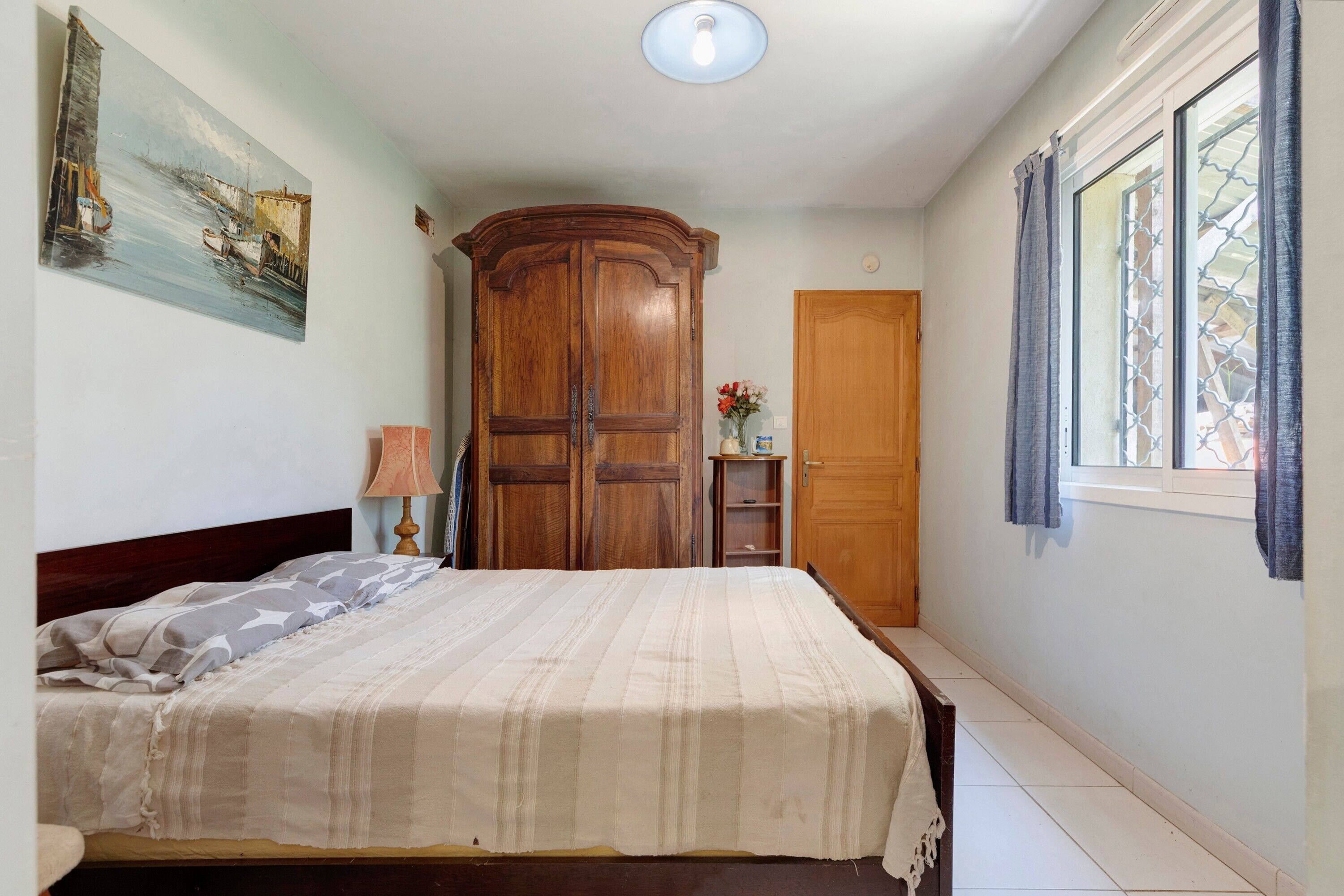 A bedroom with a bed, a wooden wardrobe, a nightstand, a lamp, a painting, a door, and a window with curtains.