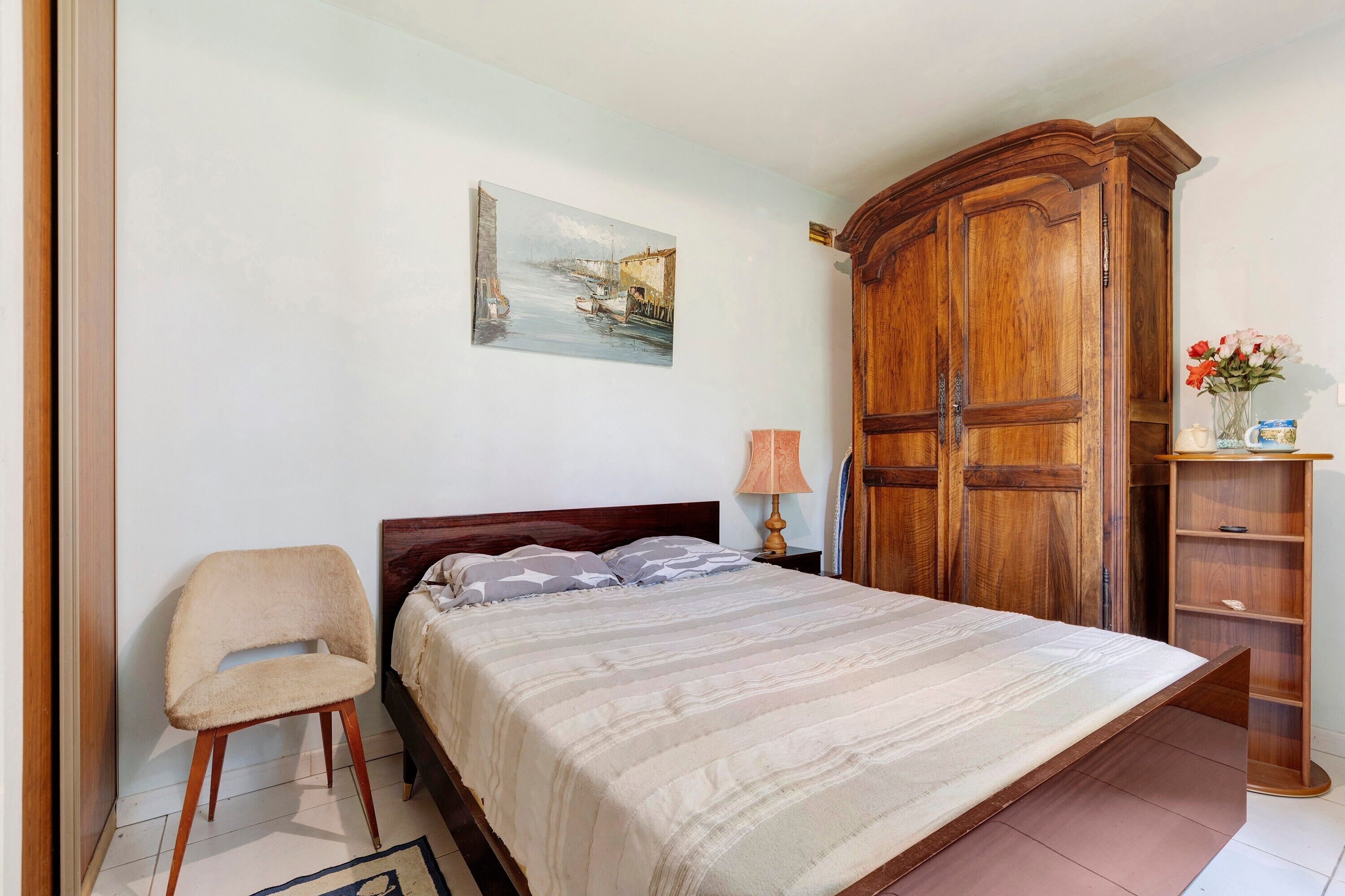 A bedroom with a bed, a chair, a wooden wardrobe, a painting on the wall, and a small shelf with flowers.