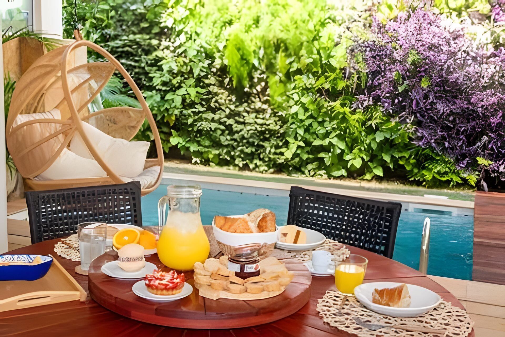 Una mesa de comedor con alimentos para el desayuno, una silla de mimbre y vista a la piscina.