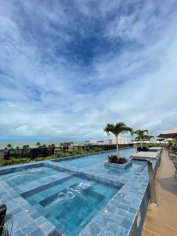 A pool area with clear blue water, palm trees, and a view of the ocean. - Luxury Apartment in HIT Cabo Branco (João Pessoa)