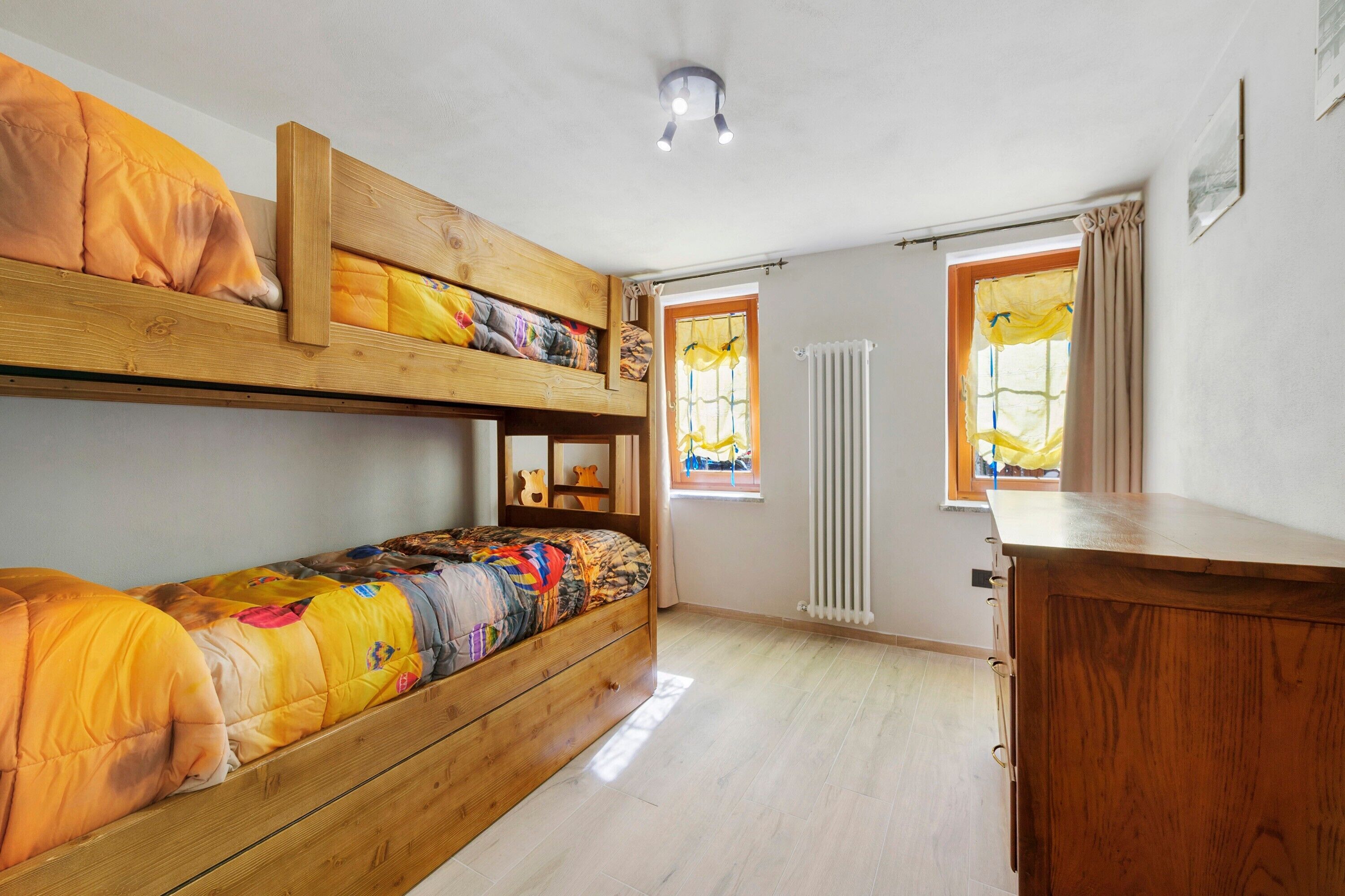 Una camera con un letto a castello, un comò in legno, una finestra con tende e un termosifone.