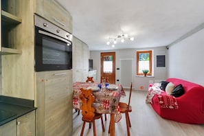 A compact living space with a dining table, wooden chairs, a red sofa, and a built-in oven. - Holiday Home 'Lueur De La Rouja' with Mountain View (Champoluc)