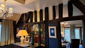 A room with exposed wooden beams, a chandelier, a desk with a lamp, and a dining area with chairs.