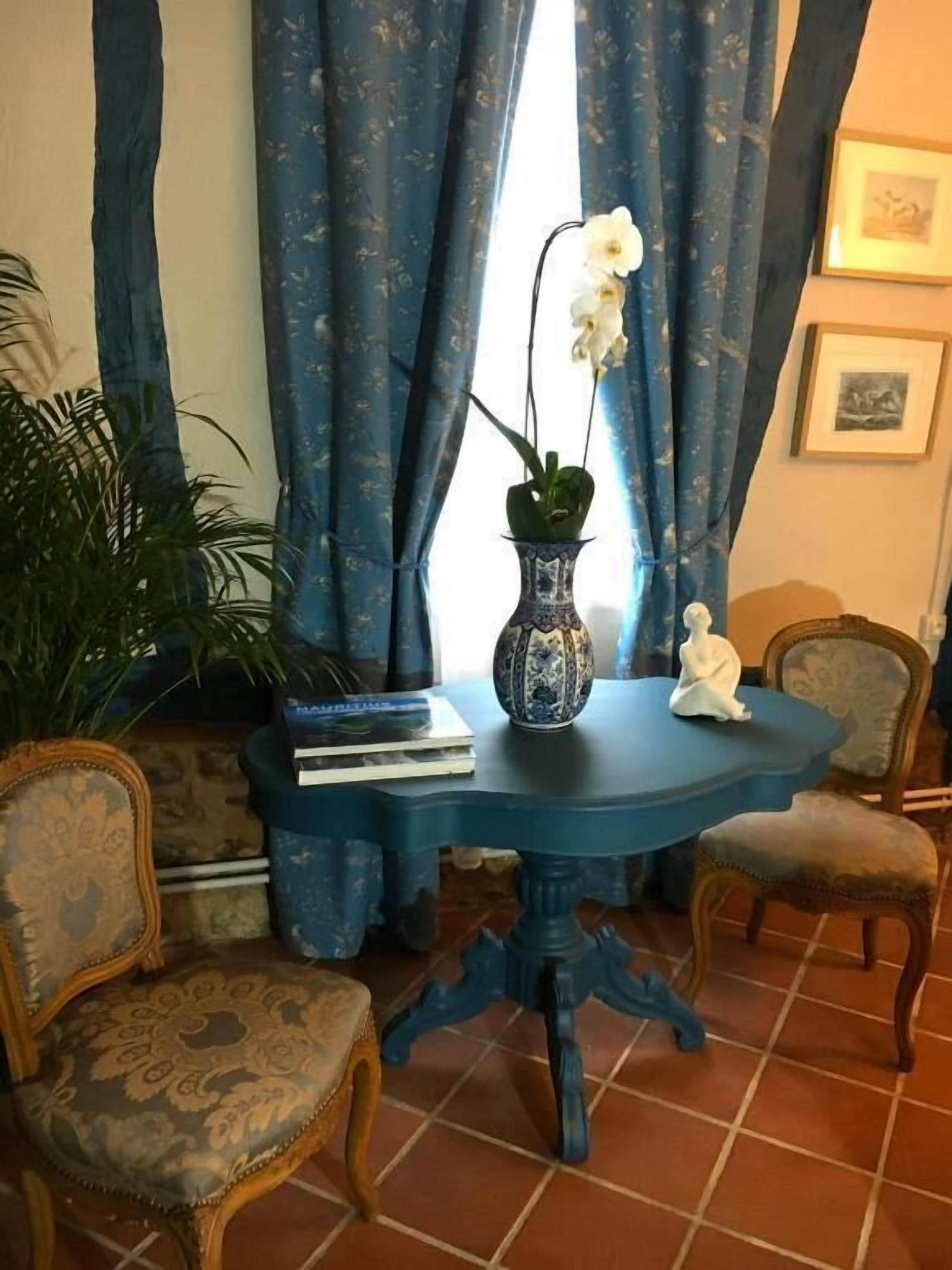Een blauwe tafel met een witte vaas en een witte beeldje, omringd door twee gestreepte stoelen, in een kamer met blauwe gordijnen en een betegelde vloer.