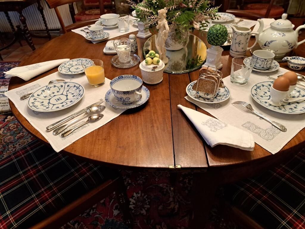 Een ronde houten tafel gedekt voor het ontbijt, met theeservies, een bloemenvaas en een decoratieve fruitschaal.