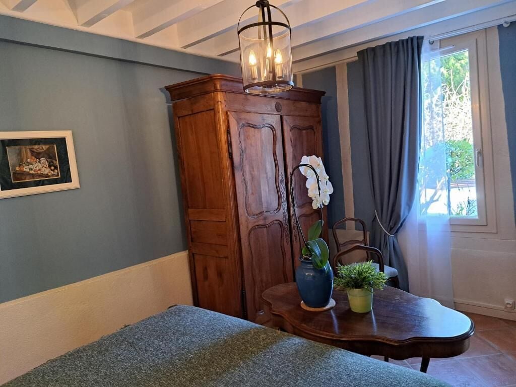 A room with a wooden cabinet, a table with a vase and plant, a framed picture on the wall, and a window with curtains.