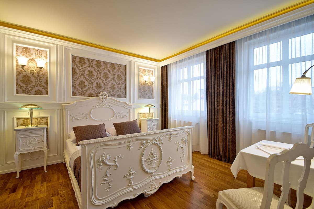 Ein Schlafzimmer mit einem großen, verzierten Bett, Nachttischen mit Lampen, einem kleinen Tisch mit Stuhl und einem großen Fenster mit Vorhängen.