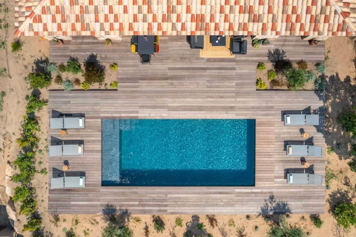 Vista aérea de una piscina rectangular con cubierta de madera, un área de estar techada con mesas y sillas, y un tejado de baldosas.