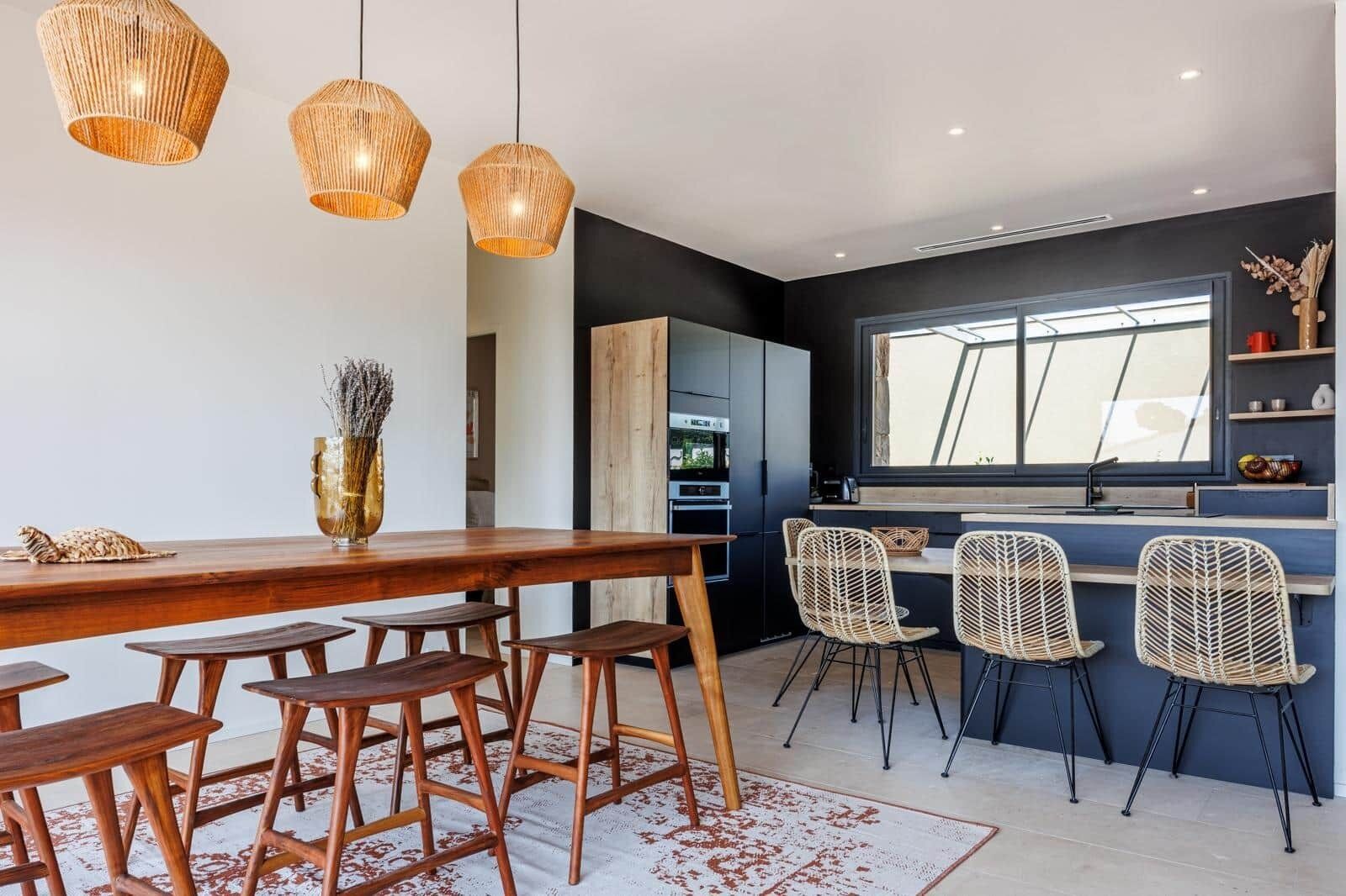 Una cocina moderna con un amplio ventanal, una mesa de comedor de madera, sillas de mimbre y lámparas colgantes.