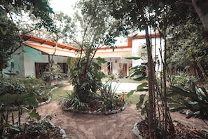 Un bâtiment traditionnel avec un toit en tuiles, entouré d’un jardin où poussent différentes espèces de plantes et bordé d’un chemin de terre.